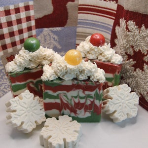 May include: Three handmade soap bars with a red, green, and white swirl design. Each bar is topped with a white whipped cream-like topping and a colorful candy. The soaps are displayed on a white surface with snowflake-shaped soaps.