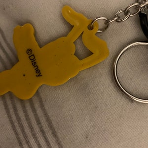 Könnte beinhalten: Gelber Plastik-Schlüsselanhänger mit einer Comicfigur und dem Text "©Disney" auf der Rückseite. Der Schlüsselanhänger ist an einem silbernen Metallring und einer Kette befestigt.