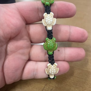 May include: A black string bracelet with four turtle-shaped beads. Two beads are green and two are white.
