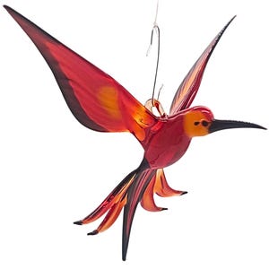 Könnte beinhalten: Ein farbenfrohes Glas-Kolibri-Ornament. Der Vogel hat einen Farbverlauf aus Rot, Orange und Gelb, mit einem langen, schwarzen Schnabel und Schwanzfedern. Eine silberne Drahtschlaufe ist zum Aufhängen angebracht. Ideal, um einen Hauch von Natur zu verleihen.