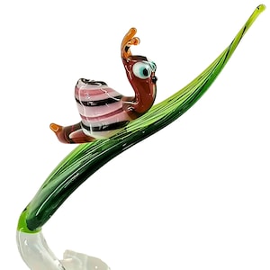 Könnte beinhalten: Eine Glasskulptur einer Schnecke mit rosa und schwarzen Streifen auf ihrer Schale, die auf einem grünen Blatt kriecht.