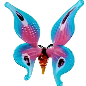 Może przedstawiać: Szklana figurka motyla z niebieskimi i różowymi skrzydłami i brązowym ciałem. Skrzydła mają białe i czarne akcenty.