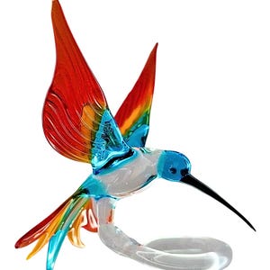 Może przedstawiać: Kolorowa figurka kolibra wykonana ze szkła z czerwonymi, żółtymi, niebieskimi i zielonymi skrzydłami. Koliber jest w locie i ma długi, czarny dziób. Figurka stoi na przezroczystej szklanej podstawie.