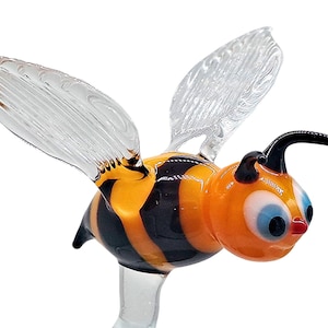 Op de afbeelding: Een glazen figuur van een bij met zwarte en gele strepen, heldere vleugels en grote blauwe ogen. De bij is in de vlucht met zijn vleugels uitgespreid.
