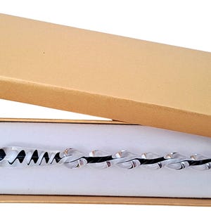 Puede incluir: Una pluma de cristal transparente y negra con diseño en espiral, colocada dentro de una caja de regalo rectangular de cartón color canela con un inserto de espuma blanca. La pluma presenta una punta afilada y un cuerpo retorcido en la caja.