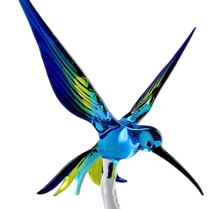 Może przedstawiać: Szklana figurka kolibra w kolorze niebieskim, zielonym i żółtym z długim czarnym dziobem. Koliber jest w locie z rozpostartymi skrzydłami.