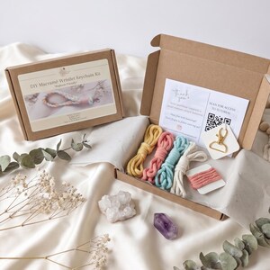 May include: A DIY macrame wristlet keychain kit in a brown box. The kit includes colorful macrame cords in yellow, pink, blue, and white, along with a gold-colored clasp. The box also contains a thank you note and a QR code for a tutorial.
