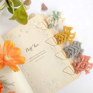 May include: Five handmade macrame bookmarks with gold metal clips. The bookmarks are in various colors, including green, white, yellow, gray, and pink. The bookmarks are hanging from a book with the text "Part Two" and a quote from Marco Polo.