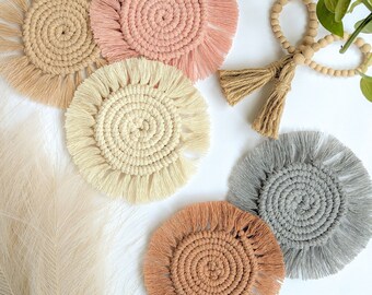 May include: Five handmade crocheted coasters with fringe in various colors, including beige, light brown, pink, and gray. The coasters are arranged on a white surface.