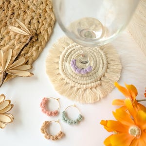 Könnte beinhalten: Drei Paar handgefertigte Creolen mit Makramee-Design. Die Ohrringe sind in den Farben Rosa, Blau und Beige gehalten. Die Ohrringe werden auf einer weißen Oberfläche mit einem gewebten Untersetzer und einem Glas präsentiert.