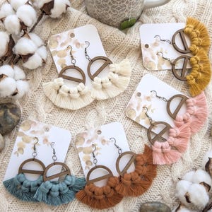 May include: Handmade macrame earrings in various colors, including cream, mustard yellow, and dusty rose. Each pair features a wooden hoop design with a fringe of knotted cotton. The earrings are displayed on white cards.