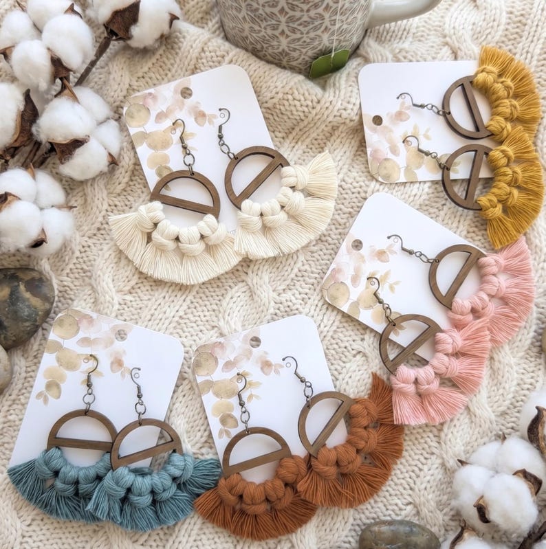 May include: Handmade macrame earrings in various colors, including cream, mustard yellow, and dusty rose. Each pair features a wooden hoop design with a fringe of knotted cotton. The earrings are displayed on white cards.