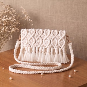 May include: A white macrame crossbody bag with a braided strap and tassel fringe. The bag features a woven design and gold-colored hardware. The bag is displayed on a wooden surface with decorative pearls and dried flowers.