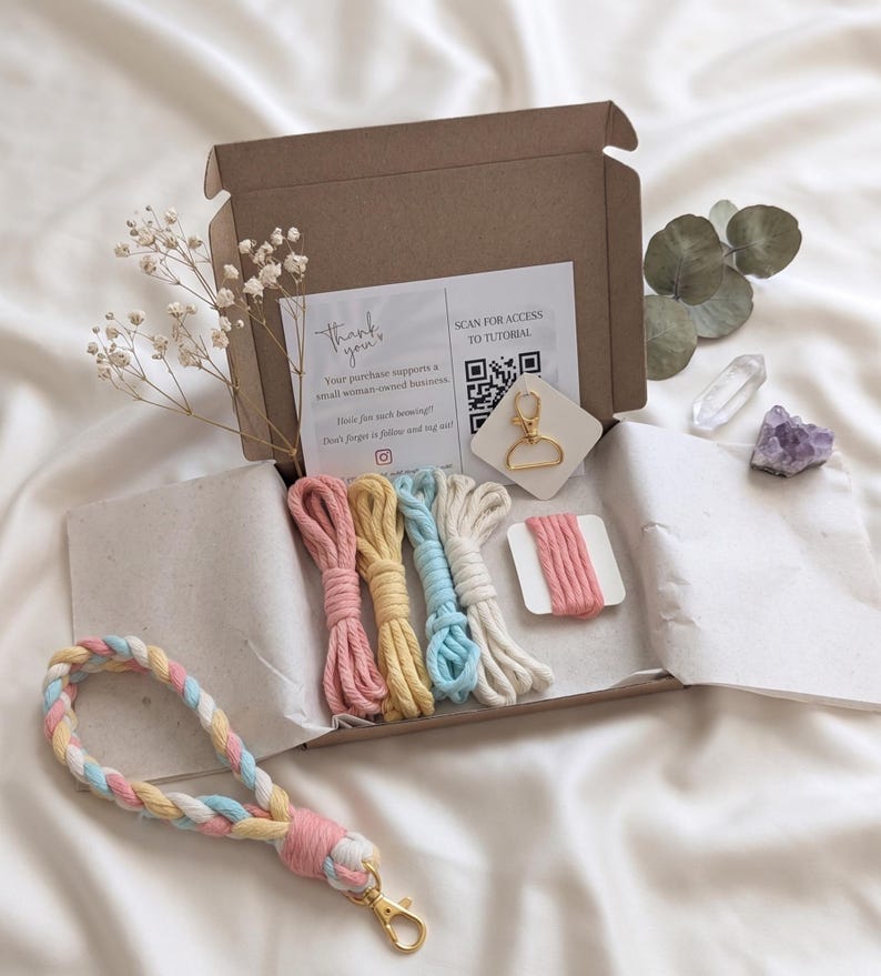 May include: An open craft box with macrame supplies, including colorful cords in pink, yellow, blue, and white. A finished keychain wristlet in pastel colors is visible. The box includes a gold-colored keychain clasp and a card with text and a QR code.