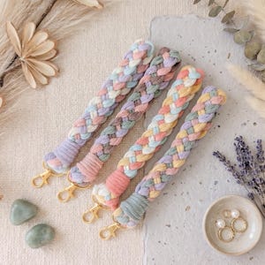 May include: Four braided wristlet keychains made of soft cotton cord in pastel color palettes including pink, blue, yellow, and lavender. Each keychain features a gold-toned metal lobster clasp at the end and a tightly wrapped cord section.