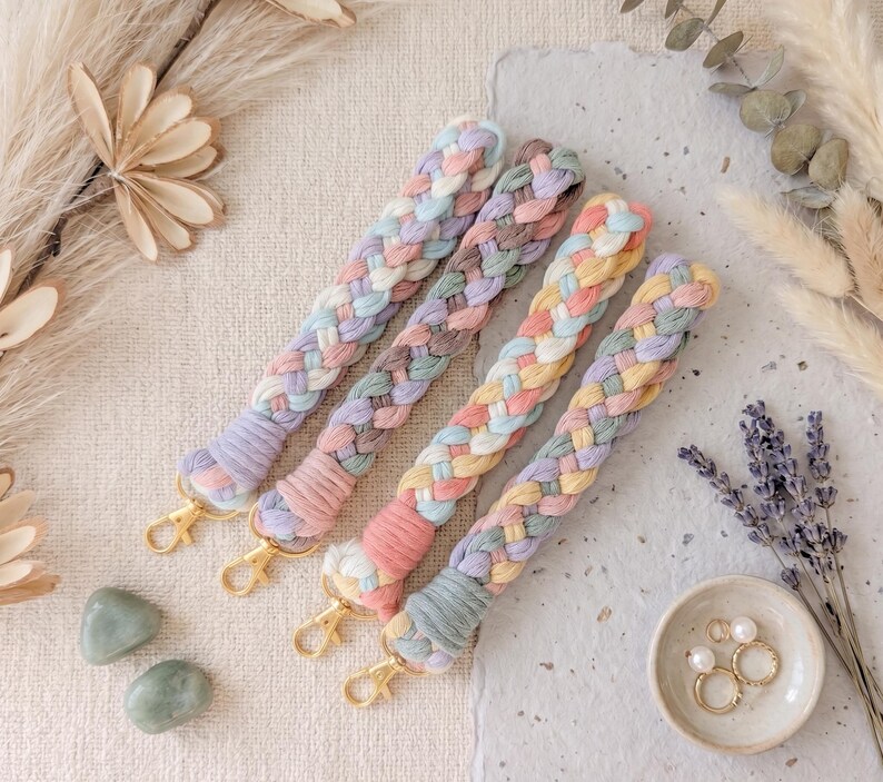 May include: Four braided wristlet keychains made of soft cotton cord in pastel color palettes including pink, blue, yellow, and lavender. Each keychain features a gold-toned metal lobster clasp at the end and a tightly wrapped cord section.
