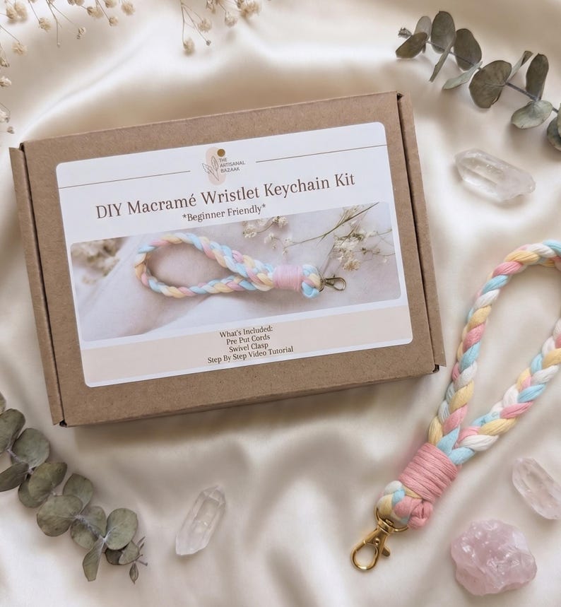 May include: A brown cardboard box labeled DIY Macram&eacute; Wristlet Keychain Kit with a pastel braided wristlet featuring pink, yellow, white, and blue cords attached to a gold swivel clasp, alongside a matching finished wristlet keychain.