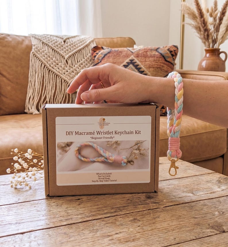 May include: A brown cardboard box labeled DIY Macram&eacute; Wristlet Keychain Kit with a picture of a braided pastel keychain. A matching braided wristlet keychain in pink, yellow, and blue with a gold-tone metal clasp is shown next to the box.