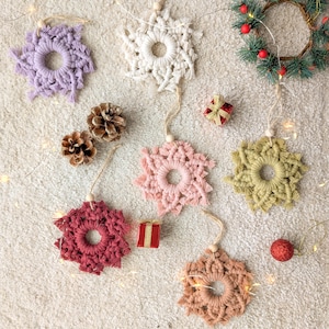 May include: A collection of seven handmade macrame snowflake ornaments in various colors, including pink, purple, red, beige, and green. The ornaments are hanging from a white surface with string and wooden beads.