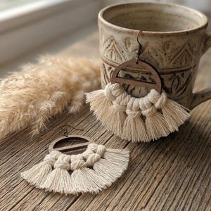 May include: Wooden and macrame earrings with a half-circle design and cream-colored tassels. The earrings are displayed on a wooden surface next to a ceramic mug. The earrings have a boho style.