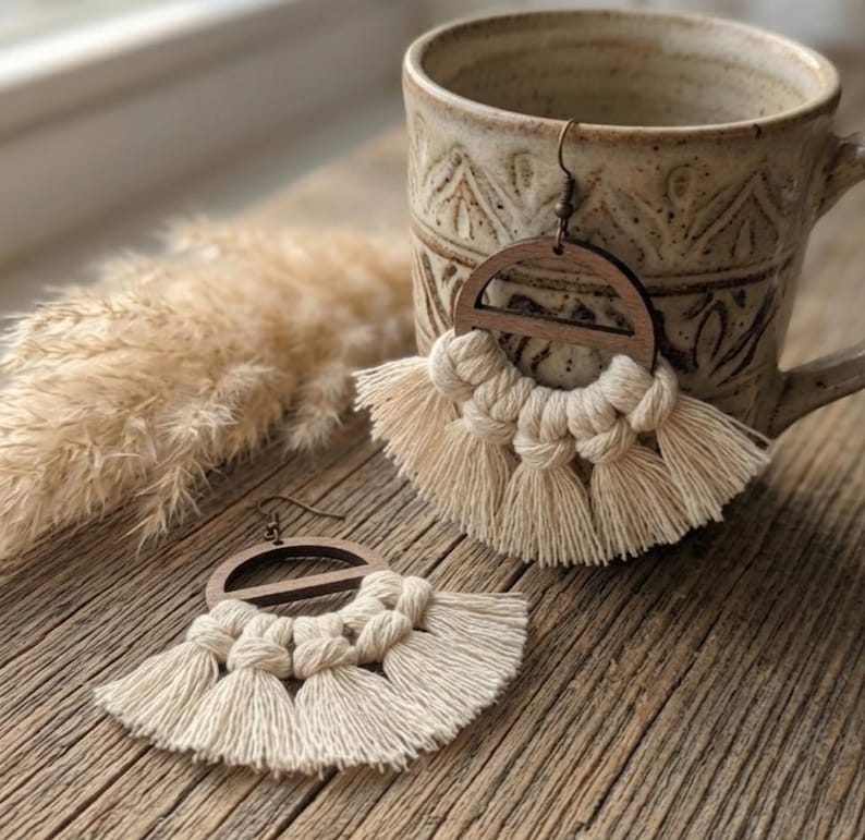 May include: Wooden and macrame earrings with a half-circle design and cream-colored tassels. The earrings are displayed on a wooden surface next to a ceramic mug. The earrings have a boho style.