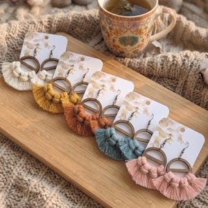 May include: A collection of handmade macrame earrings in various colors, including cream, yellow, orange, teal, and pink. Each earring features a wooden semi-circle design and a fringe tassel. The earrings are displayed on white cards.
