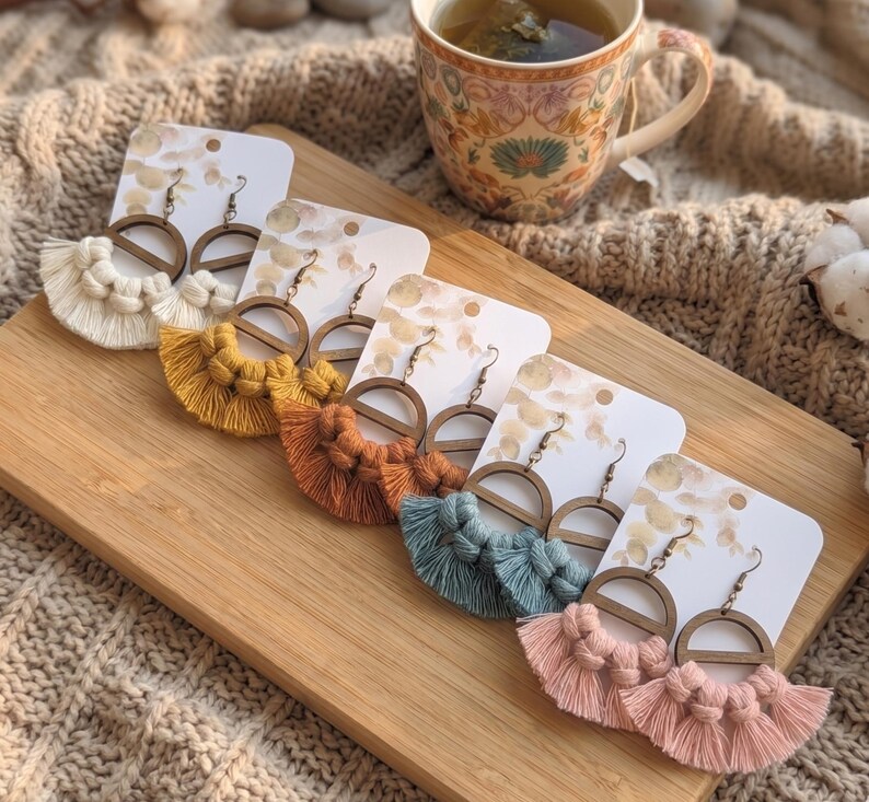 May include: A collection of handmade macrame earrings in various colors, including cream, yellow, orange, teal, and pink. Each earring features a wooden semi-circle design and a fringe tassel. The earrings are displayed on white cards.
