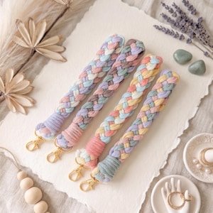 May include: Four pastel-colored braided wrist straps with gold-colored clasps. The straps are in shades of pink, blue, purple, and yellow. The background is a textured off-white surface with decorative elements.