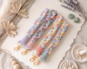 May include: Four pastel-colored braided wrist straps with gold-colored clasps. The straps are in shades of pink, blue, purple, and yellow. The background is a textured off-white surface with decorative elements.