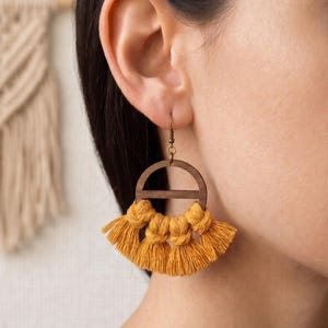 May include: Mustard yellow tassel earrings with a wooden semi-circle design. The earrings have a brass hook and are shown on a person's ear. The tassels are knotted at the top and fan out at the bottom.