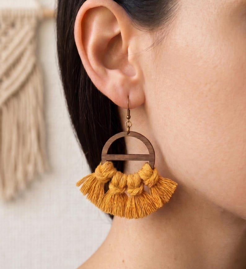 May include: Mustard yellow tassel earrings with a wooden semi-circle design. The earrings have a brass hook and are shown on a person's ear. The tassels are knotted at the top and fan out at the bottom.