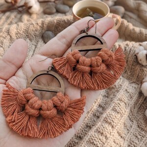 May include: Handmade macrame earrings with a wooden semi-circle design. The earrings feature a burnt orange color with knotted details and fringe. The earrings are displayed on a hand, showcasing the intricate craftsmanship and bohemian style.