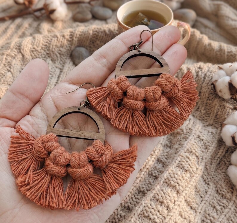 May include: Handmade macrame earrings with a wooden semi-circle design. The earrings feature a burnt orange color with knotted details and fringe. The earrings are displayed on a hand, showcasing the intricate craftsmanship and bohemian style.