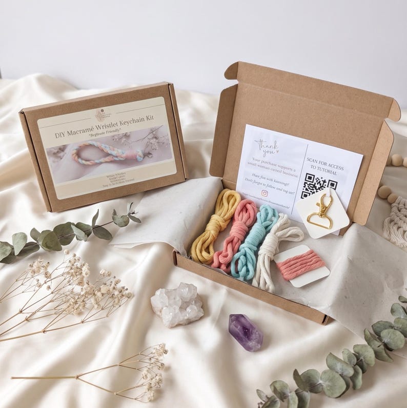 May include: A DIY macrame wristlet keychain kit in a brown box. The kit includes colorful macrame cords in yellow, pink, blue, and white, along with a gold-colored clasp. The box also contains a thank you note and a QR code for a tutorial.