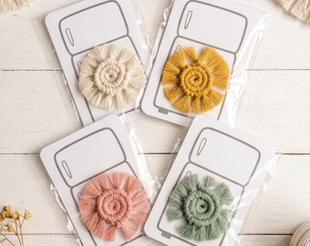 May include: Four handmade macrame coasters in cream, mustard yellow, dusty rose, and sage green, each packaged on a card with a refrigerator illustration. The coasters have a spiral center with fringed edges, perfect for home decor.