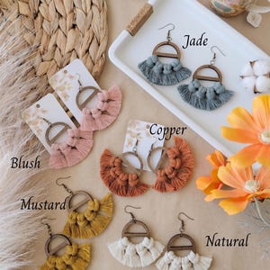 May include: A collection of handmade macrame earrings in various colors, including blush, copper, mustard, jade, and natural. Each pair features a wooden semi-circle and a fringe design. The earrings are displayed on cards.