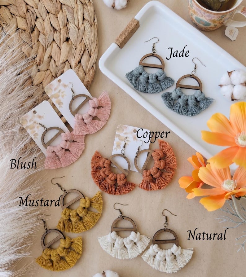 May include: A collection of handmade macrame earrings in various colors, including blush, copper, mustard, jade, and natural. Each pair features a wooden semi-circle and a fringe design. The earrings are displayed on cards.
