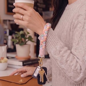 May include: A braided wristlet keychain featuring soft pastel colors including pink, blue, and yellow. The wristlet is attached to a gold-toned metal lobster clasp that holds a black car key fob and a set of silver and brass metal keys.