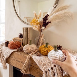 May include: Four handmade macrame pumpkins in rust, cream, mustard yellow, and white, each featuring a felt stem. The pumpkins are arranged on a textured beige runner alongside a glass vase with dried flowers, wooden beads, and pinecones.
