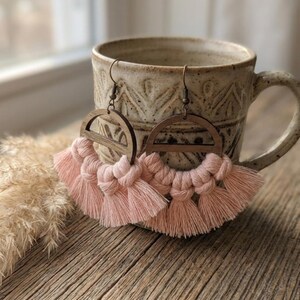 May include: A pair of handmade macrame earrings. The earrings feature a wooden semi-circle design at the top, with a fringe of soft pink tassels below. The earrings are hanging in front of a ceramic mug on a wooden surface.