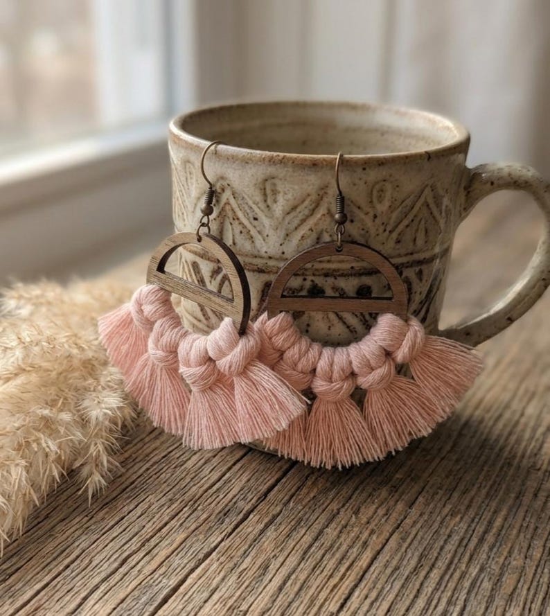 May include: A pair of handmade macrame earrings. The earrings feature a wooden semi-circle design at the top, with a fringe of soft pink tassels below. The earrings are hanging in front of a ceramic mug on a wooden surface.