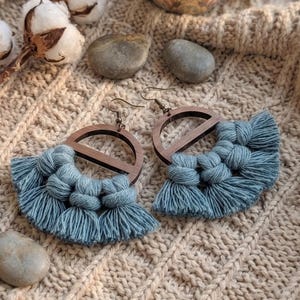 May include: A pair of handmade earrings featuring a wooden semi-circle design with a blue macrame fringe. The earrings have a hook closure and are displayed on a textured, beige surface. Cotton buds and small stones are also visible.