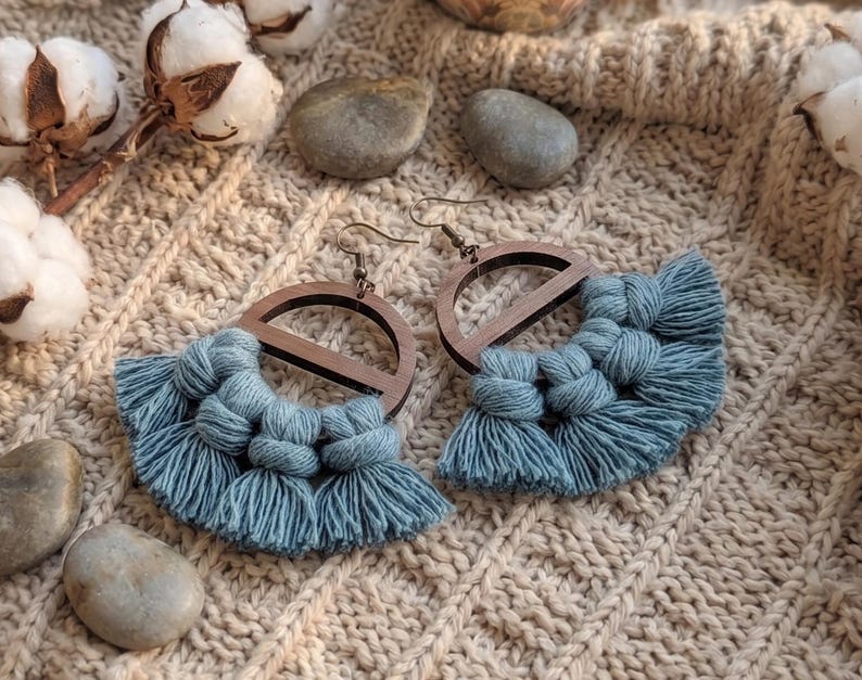May include: A pair of handmade earrings featuring a wooden semi-circle design with a blue macrame fringe. The earrings have a hook closure and are displayed on a textured, beige surface. Cotton buds and small stones are also visible.