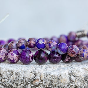 May include: Two strands of purple and white marbled gemstone beads. The beads are round and have a speckled pattern. The beads are strung on a silver cord.