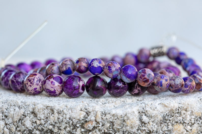 May include: Two strands of purple and white marbled gemstone beads. The beads are round and have a speckled pattern. The beads are strung on a silver cord.