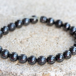 May include: A black hematite beaded bracelet with a shiny finish. The bracelet is made of round beads and is strung on a clear elastic cord.