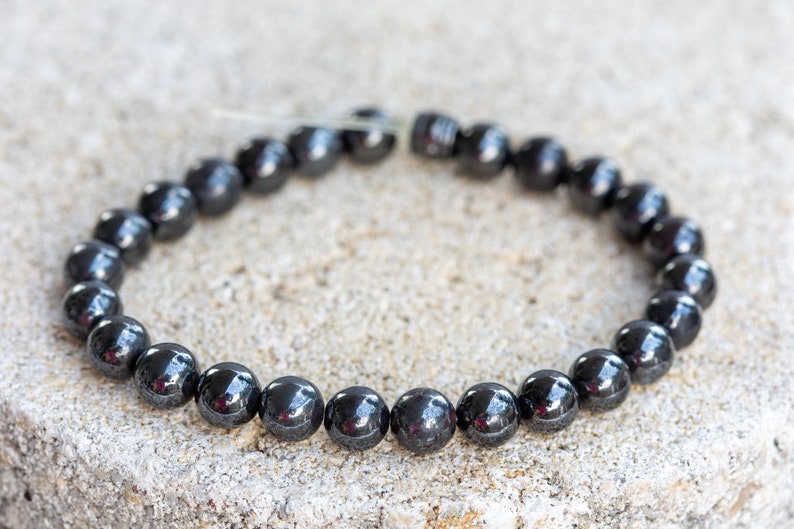 May include: A black hematite beaded bracelet with a shiny finish. The bracelet is made of round beads and is strung on a clear elastic cord.