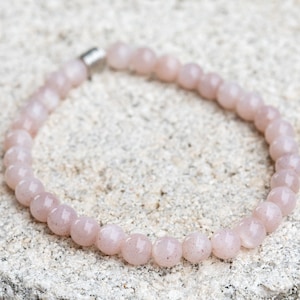 May include: A light pink beaded bracelet with a silver clasp. The bracelet is made of round, polished beads. The bracelet is resting on a textured, light-colored surface. Jewelry.
