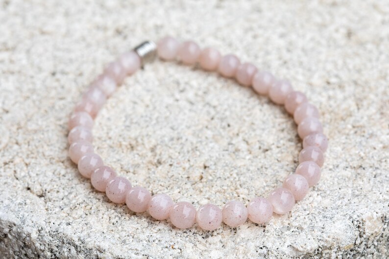 May include: A light pink beaded bracelet with a silver clasp. The bracelet is made of round, polished beads. The bracelet is resting on a textured, light-colored surface. Jewelry.
