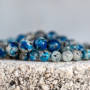 May include: A beaded bracelet made of blue and white kyanite stones. The bracelet is made of round beads and is stacked on top of another bracelet.